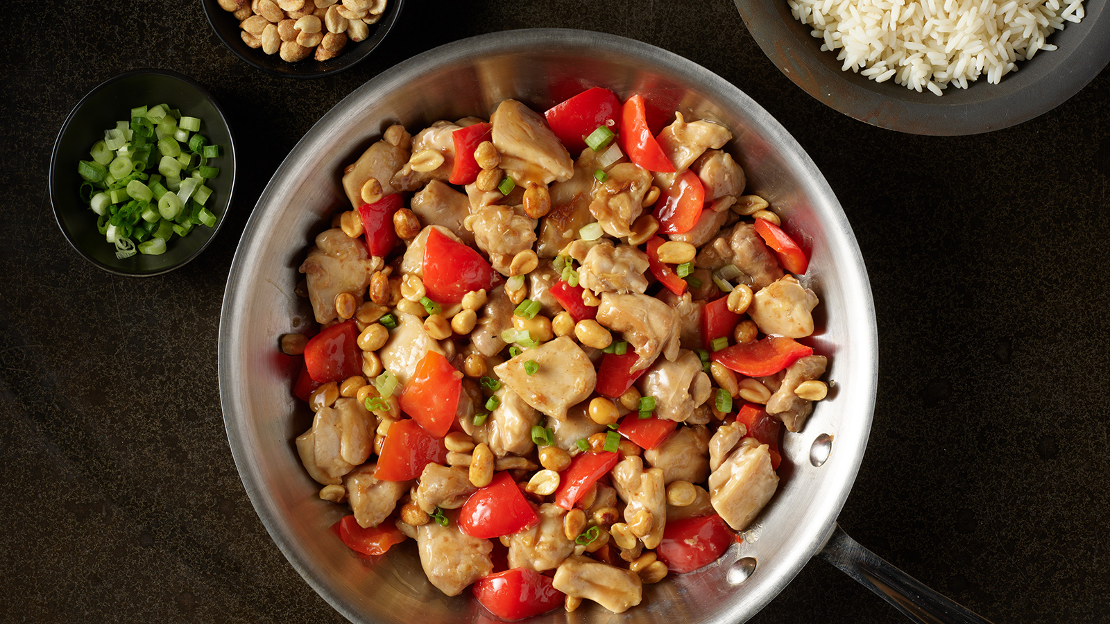 Pan of spicy peanut chicken with small bowls of sliced green onions, peanuts and white rice.