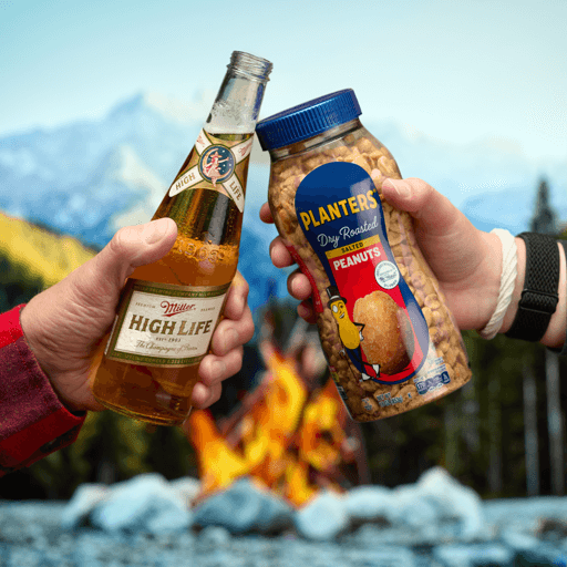Two people cheers’ a miler high life and a container of planters bar nut mix with a mountain background