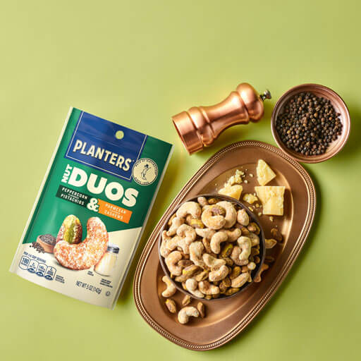 Promotional photo of planters duos peppercorn pistachios and parmesan cashews with a charcuterie board on a green background