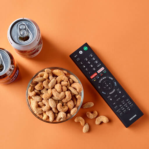 Promotional photo of planters duos ranch almonds and buffalo cashews on an orange background with cans and a tv remote